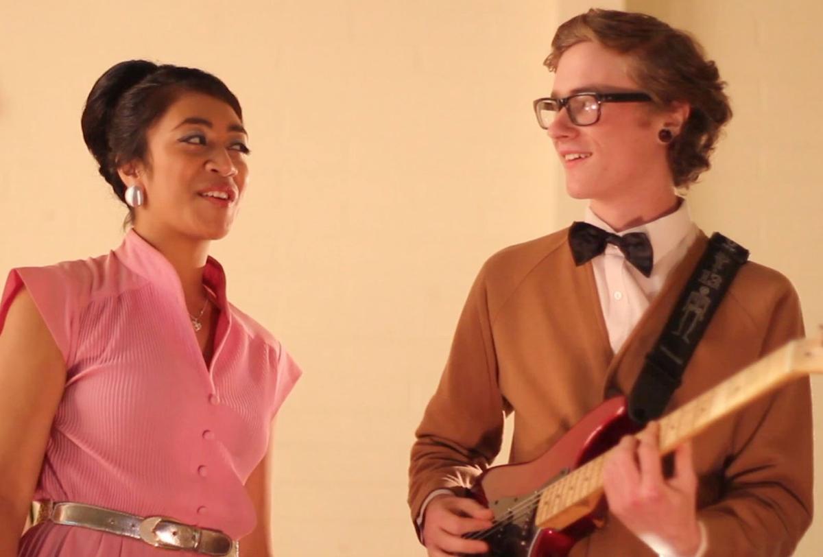 A femme person in a pink dress smiles whilst singing at a pākehā guy who is playing a gutair. They are standing next to each other on what feels like a stage with a plain background.